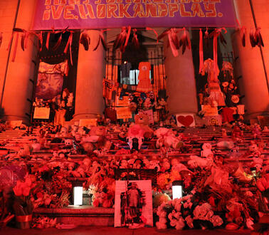 Das Bild zeigt Treppen vor einem Gebäude in roter Beleuchtung mit Kerzen, Bildern, Blumen und Spielsachen.