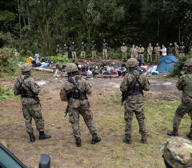 Das Bild zeigt mehrere Soldaten, die eine Gruppe von Menschen bewachen, die vor ihnen auf dem Boden kauern.
