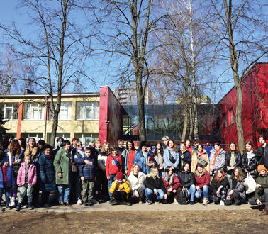 Kinder einer Schulklasse haben sich zusammen mit ihren Lehrerinnen zum Foto aufgestellt, hinter ihnen stehen Bäume und Schulgebäude.Schüler_innen und Lehrerinnen aus Charkiw vor der Migrationsbehörde in Vilnius aufgestellt.