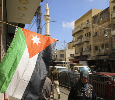 Aufnahme einer Straße. Links im Bild befindet sich eine jordanische Flagge. Auf der rechten Seite läuft eine Frau mit Hijab.