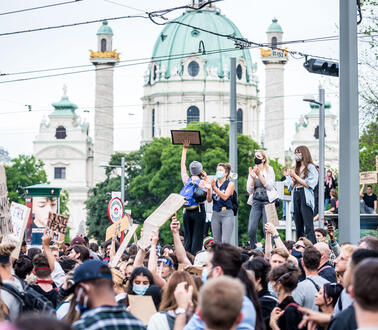 Foto einer Menschenmenge mit Schildern und Masken. Im Hintergrund befindet sich die Karlskirche.