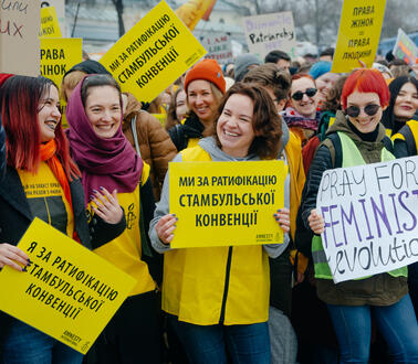 Das Foto zeigt eine große Menschenmenge. Im Vordergrund stehen mehrere junge Frauen, die Protestschilder hochhalten und sich anlächeln.