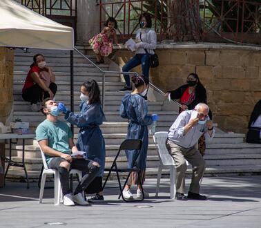 Auf Stühlen bei einem Pavillion werden zwei Personen von medizinischem Personal in Schutzkleidung auf Corona getestet.