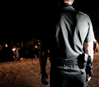 Das Bild zeigt eine Situation in der Nacht: Im Vordergrund einen Polizisten von hinten, im Hintergrund eine Gruppe von Personen, die am Boden sitzt.