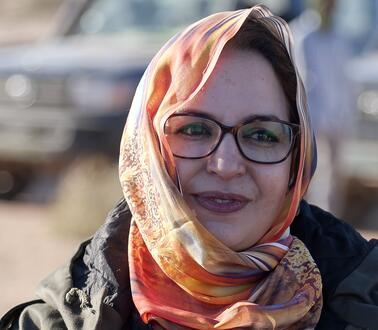 Eine junge Frau trägt ein buntes Kopftuch und eine Brille, und lächelt.