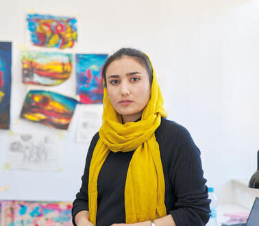 Die afghanische Künstlerin Nabila Horakhsh steht in ihrem Hamburger Atelier, sie trägt Jeans, einen Silberarmreif und Ringe an den Fingern, sowie ein Kopftuch, sie lehnt an einem Schreibtisch, auf dem ein Laptop steht und ein offener Pappkarton mit Joghurtbechern und Gläsern, die Farbe enthalten, eine Wasserflasche steht auf dem Tisch, im Hintergrund eine  farbverschmierte Leinwand und Bilder auf Papier, die an die Wand geklebt worden sind.