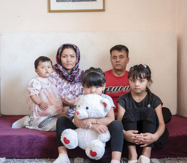 Eine afghanische Familie sitzt auf einer Matratze mit Frotteebezug, die auf dem Boden liegt in einem Zimmer, an die Eand hinter ihnen ist eine weitere Matratze gelehnt, die Mutter mit Kopftuch hält ein Kleinkind halb auf dem Schoß, neben ihr sitzt der Vater, vor ihnen ihre zwei Töchter auf der Kante der Matratze, eine versteckt ihr Gesicht halb hinter einem großen Teddybären, die andere hat ihre Beine angezogen und ihre Arme darum geschlungen.