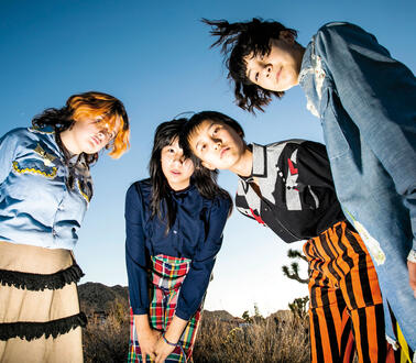 Vier Mädchen, drei von ihnen tragen Röcke und Hemden mit Kragen stehen draußen in einer hügeligen Landschaft. 