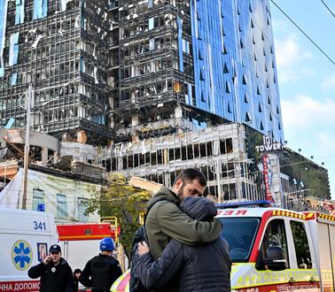 Das Bild zeigt im Vordergrund zwei Menschen, die sich umarmen, im Hintergrund ein zerstörtes Gebäude, 