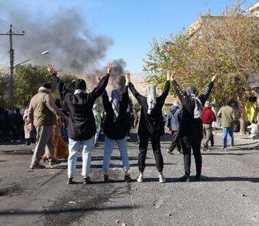Vier Frauen stehen mit dem Rücken zur Kamera auf einer Straße. Sie heben ihre Hände in die Luft und zeigen mit beiden Händen das Victory-Zeichen. Weitere Menschen demonstrieren auf der Straße. Im Hintergund steigt Rauch auf.