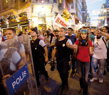 Menschen stehen dicht gedrängt in einer Fußgängerzone, sie demonstrieren, eine Frau trägt eine Atemschutzmaske um den Hals und eine Taucherbrille, die sie in die Stirn geschoben hat, neben und vor ihr laufen Sicherheitskräfte und türkische Polizisten.