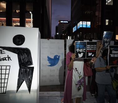 Demonstrant*innen stehen mit Plakaten im Dunkeln auf einer Straße, auf ihren Schildern steht "Shout No Hate" und auf anderen sind japanische Schriftzeichen und das Logo von Twitter abgebildet.