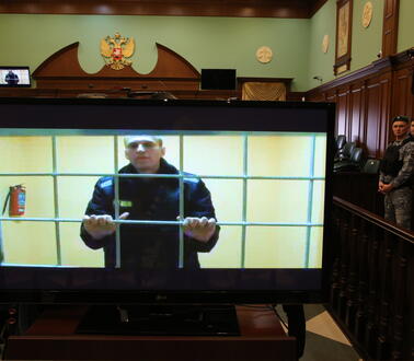 Das Bild zeigt einen TV-Bildschirm, der in einem Gerichtssaal steht. Darauf zu sehen, ein Mann hinter Gittern.
