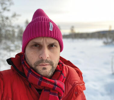 Ein Mann mit Dreitagebrat trägt Mütze und Schal in einer verschneiten Landschaft.