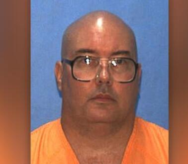 Porträtfoto von Donald Dillbeck, dessen Kopf kahlrasiert ist und der eine Brille und Gefängniskleidung trägt.