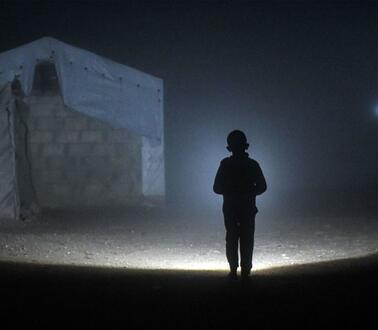 Das Foto zeigt die Silhouette eines Jungen von hinten bei Nacht und Nebel. Seine Taschenlampe wirft einen großen Lichtkegel voraus. Links von dem Jungen steht ein großes Zelt, das mit Mauern verstärkt ist.