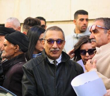 Das Foto zeigt Ihsane El Kadi, der einen Anzug mit Krawatte trägt und eine Sonnenbrille. Er steht umgeben von mehreren Personen vor einem Gebäude und blickt in die Kamera.
