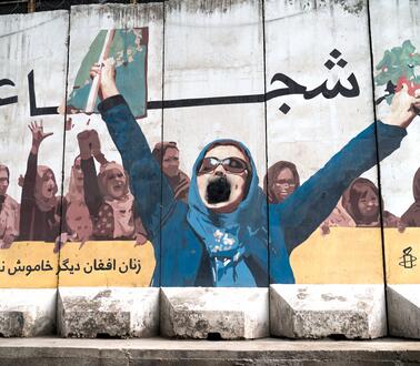 Das Bild zeigt eine Mauer mit einem Grafitti, darauf zusehen eine Frau, die die Hände die Höhe hebt