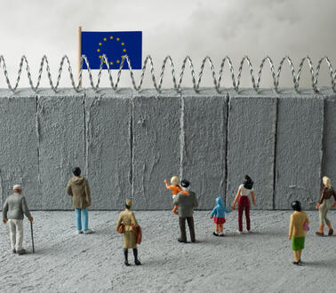 Das Foto zeigt eine Installation mit Spielzeugfiguren. Sie stehen vor einer hohen Mauer, die mit Stacheldraht versehen ist. Dahinter weht die Flagge der Europäischen Union.