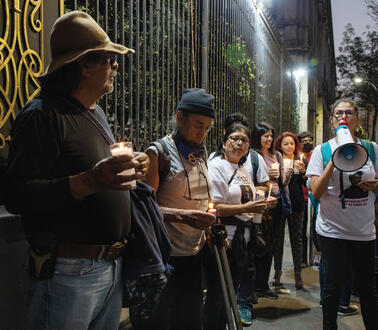 Demonstrant*innen auf einer Straße stehen vor einem verzierten schmiedeeisernen Zaun, eine Frau spricht in ein Megafon. Ein Mann mit Hut und Bart hält ein Getränk in der Hand und hört ihr zu.