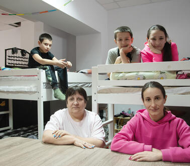 Eine Familie in einem Zimmer einer Flüchtlingsunterkunft, zwei Hochbetten, darauf liegend und sitzend drei Kinder, ein Junge und zwei Mädchen, vor den Hochbetten am Tisch zwei Frauen, sitzend.