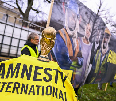 Das Bild zeigt ein Amnesty-Banner, darauf ein Pokal und dahinter ein großes Plakat mit Menschen in Arbeitskleidung