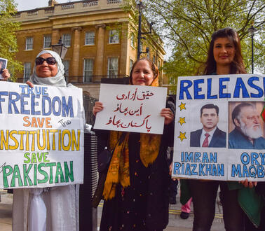 Mehrere Personen stehen vor einem Gebäude und halten Plakate hoch. Auf einem steht: "Release Imran Riaz Khan".