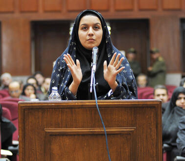 Eine junge Frau mit Kopftuch steht in einem Gerichtssaal vor einem Redepult und gestikuliert mit ihren beiden Händen.