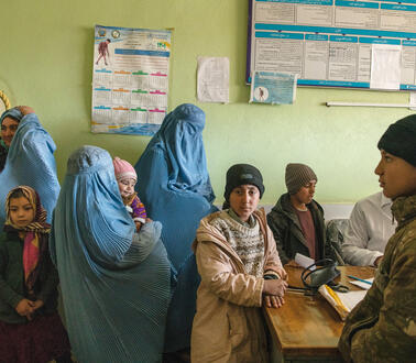 Afghanische Frauen und Kinder in traditioneller Kleidung warten in einem Zimmer eines Krankenhauses, an einem Schreibtisch sitzt ein Arzt, auf dem Tisch liegen Papiere und ein Stetoskop.
