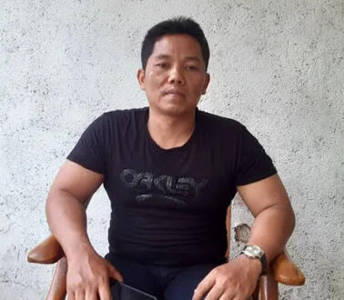 Heri Budiawan trägt ein T-Shirt und sitzt auf einem Stuhl mit beiden Armen auf den Lehnen und blickt ernst in die Kamera. 