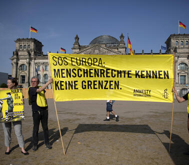 Vier Menschen vor dem Reichstagsgebäude, dem Sitz des Deutschen Bundestag. Zwei davon halten ein großes gelbes Amnesty-Transparent hoch, auf dem steht: SOS Europa – Menschenrecht kennen keine Grenzen. 