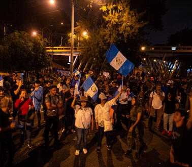 Das Foto zeigt eine große Gruppe Demonstrierender bei Nacht auf einer Straße. Einige von ihnen schwenken die Flagge Guatemalas.