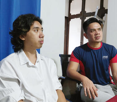 Zwei philippinische Männer sitzen nebeneinander in einem Zimmer. Einer trgät Hemd und sein gewelltes Haar bis fast zur Schulter. Der andere trägt das Haar kurz nach hinten gegelt und ein T-Shirt.