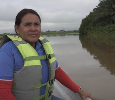 Ein Frau mit Rettungsweste sitzt in einem kleinen Boot und fährt an der Nähe des Ufers auf einem Fluss.