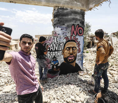 Junge Männer stehen inmitten der Ruine eines zerbombten Hauses, einer von ihnen malt ein Grafitti-Bild an einen Pfeiler, andere stehen um ihn herum, ein anderer macht ein Selfie mit seinem Handy.