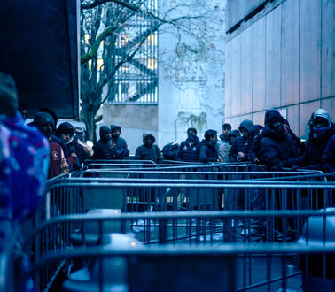Das Bild zeigt Absperrgitter, mehrere Menschen stellen sich in einer Reihe an und warten