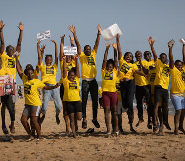 Das Bild zeigt viele junge Menschen mit gelben T-Shirts an einem Strand, sie reißen die Arme in die Luft, viele lachen