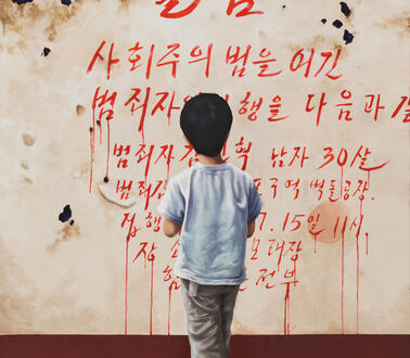 Ein kleiner Junge steht vor einer Wand, auf die koreanische Schriftzeichen in roter Farbe gemalt sind, die nach unten hin verlaufen wie Blut. 