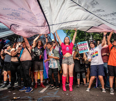 Mehrere Personen stehen auf einer Straße nebeneinander und breiten über ihren Köpfen eine große Transgender-Pride-Fahne aus.