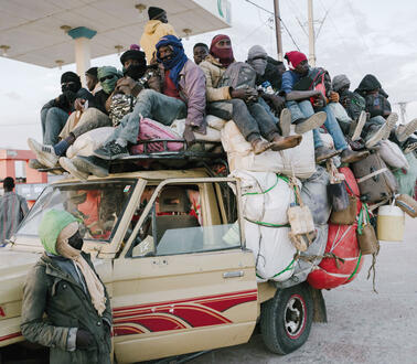Ein Allradwagen, behangen mit Gepäck, auf dem Dachgepäckträger sitzen sehr viele Menschen, die teilweise Mützen oder Masken und Sonnebrillen tragen.