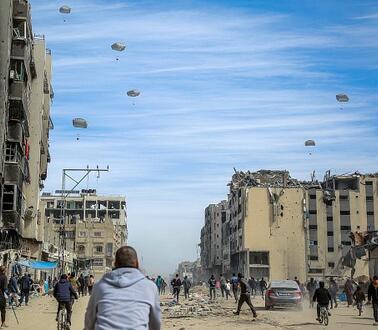 Das Bild zeigt im Vordergrund Menschen inmitten zerstörter Häuser, im Hintergrund am Himmel erkennt man mehrere Fallschirme