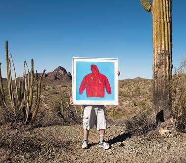 Ein Mann steht in einer Wüste, Kakteen um ihn herum, er trägt kurze Hosen und Laufschuhe und hält ein gerahmtes Bild von einem Foto eines Hoodies vor seinem Oberkörper.