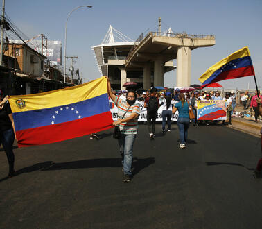 Das Bild zeigt viele Menschen auf einer breiten Straße, manchen halten die Nationalflagge Venezuelas in der Hand