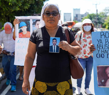 Das Bild zeigt eine Frau, die das Foto eines Mannes in der Hand halt. Hinter ihr stehen viele Menschen mit Protestplakaten