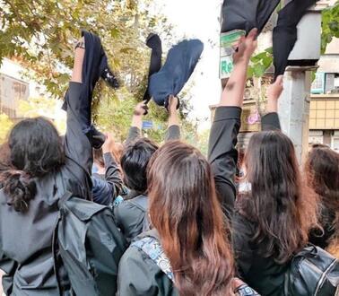 Das Bild zeigt mehrere junge Frauen von hinten, die schwarze Kopftücher in die Luft werfen