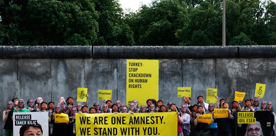Eine Gruppe von Amnesty-Aktivistinnen und Aktivisten steht vor der Berliner Mauer, hält Banner und Schilder hoch, mit Masken von İdil Eser und Taner Kılıç