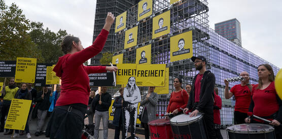 Eine Samba-Gruppe mit Trommeln neben Menschen mit Protestschildern vor einer Installation aus Wassertanks