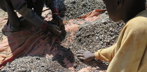 Zwei Kinder hocken auf dem Boden und wühlen mit den Händen durch Erde und Steine, die auf einer Plane ausgebreitet sind