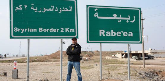 Mohammed Khamis steht vor zwei grünen Straßenschildern, auf denen "Syrian Border 2Km" und "Rabe'ea" auf Arabisch und Englisch steht