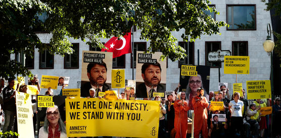 Menschen vor weißem Gebäude mit Türkei-Flagge, die gelbe Amnesty-Banner sowie Schilder mit der Forderung "Release İdil Eser and Taner Kılıç" hochhalten 